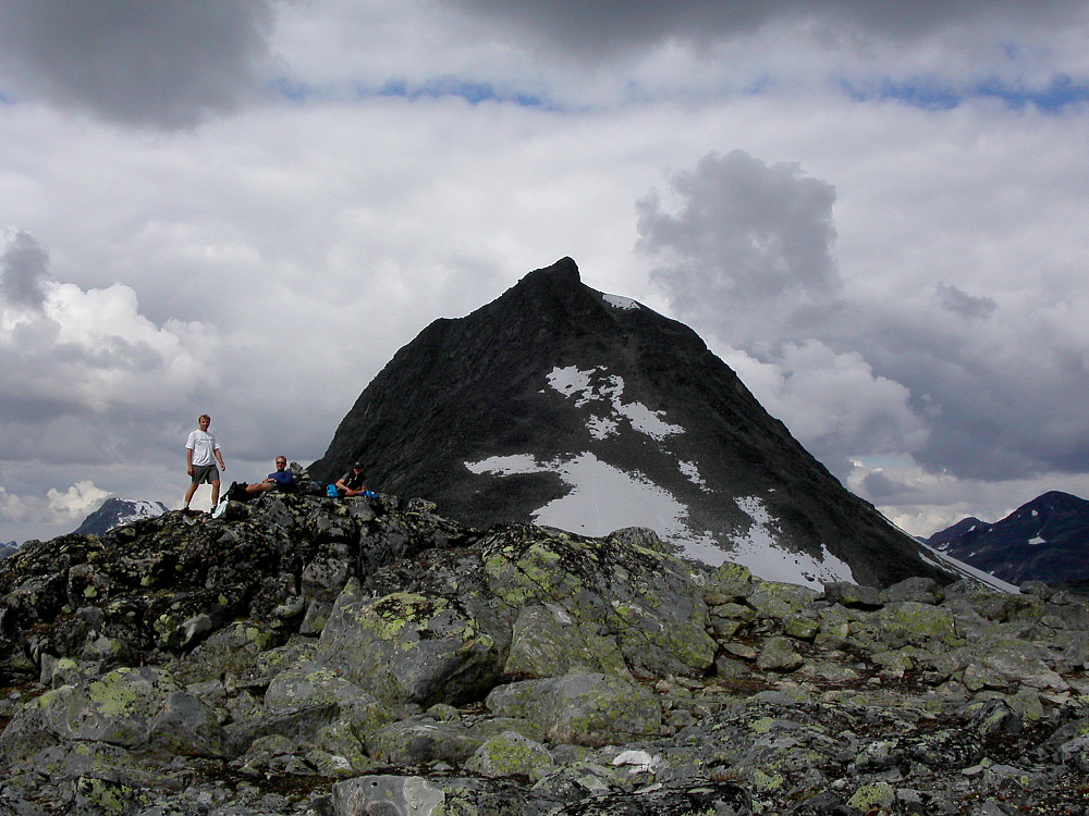 01.08.2004 - Vi tar en pause på toppen Sør for Mjølkedalstinden (1843). Selve Mjølkedalstinden (2138) dominerer bakgrunnen. Våre spor kan skimtes i den store snøfonna nederst rett under toppen.