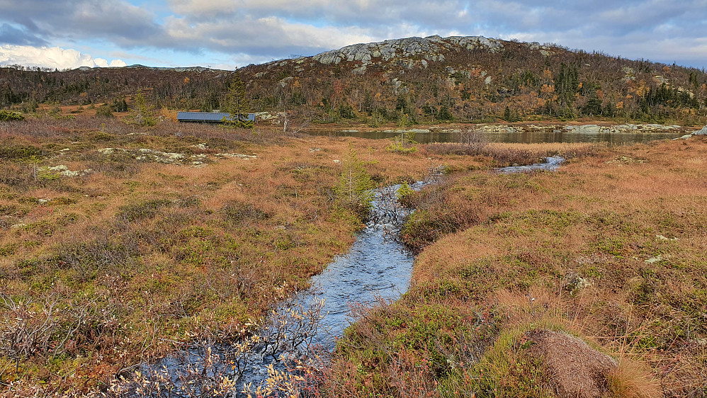 Den overfylte bekken som renner fra Store til Vesle Rustadvatn. Bygningen til venstre ligger langs vestbredden av Store Rustadvatn. Rustadstølfjellet (1090) er bak og litt til høyre.